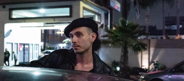 Lefty Wingate viewed from over the hood of a car there is a sign in the background. its night, he's wearing a baret.