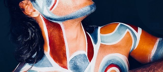 Ruely with his eyes closed and half naked covered in grey and white and red paint in swirling patterns.